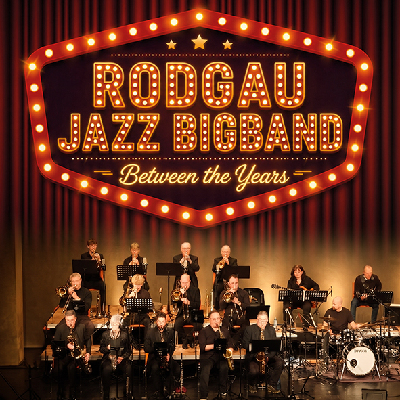 Bigband auf einer Bühne vor einem roten Vorhang, darüber ein Leuchtreklameschild mit dem Schriftzug Rodgau Jazz Bigband Between the years