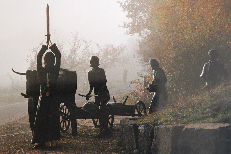 Skulptur eines keltischen Trauerzugs mit Stier, der einen Katafalk zieht, Trauernden und im Vordergrund ein Priester, der ein Schwert erhebt und ein goldenes Medaillon um den Hals trägt.