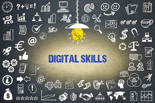 Schwarze Tafel mit blauer Schrift 'DIGITAL SKILLS' und zahlreichen weißen Symbolen zu digitalen und wirtschaftlichen Themen, darüber eine Lampe mit gelbem Papier als Glühbirne