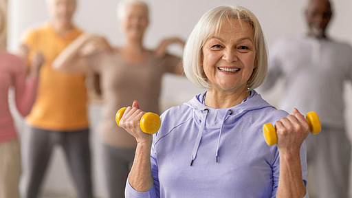 Seniorin mit gelben Hanteln im Hintergrund weitere Senioren beim Sport.