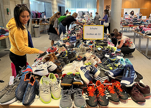 Schuhe in Größe 33 auf einem Tisch im Foyer der Kulturhalle beim Basar 2024, im Hintergrund einkaufende Frauen.