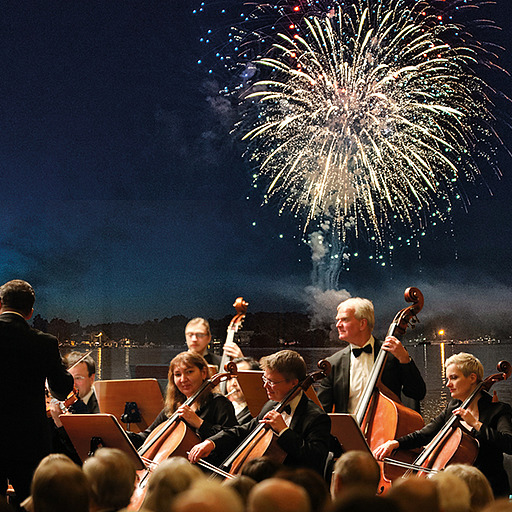 Streichorchester vor nächtlicher Flusskulisse mit Feuerwerk am Himmel.
