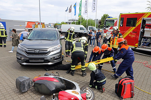 Kinder mit Bergewerkzeug an einem Unfallauto, im Vordergrund ein Motorroller am Boden.