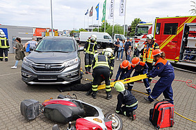 Kinder mit Bergewerkzeug an einem Unfallauto, im Vordergrund ein Motorroller am Boden.